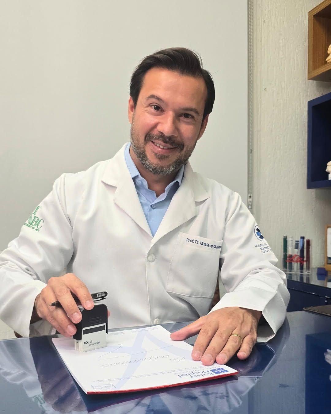 Dr. Luiz Gustavo de Quadros de jaleco, sorrindo, em seu consultório